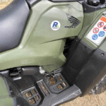 Close-up of olive-green Honda ATV body with seat, step rails, and a safety/warning label on the console panel.