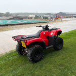 Red ATV parked on a grassy verge with an industrial complex and equipment in the background