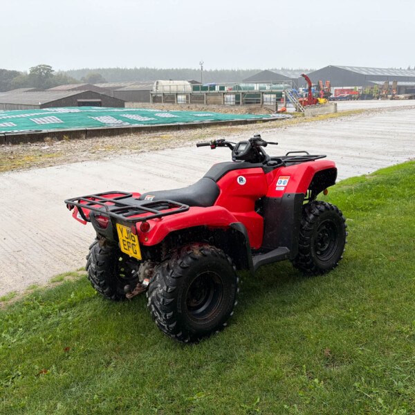 Red ATV parked on a grassy verge with an industrial complex and equipment in the background