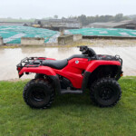 Red ATV parked on a grassy patch with a rainy, industrial yard and tarp-covered structures in the background.