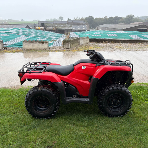 Red ATV parked on a grassy patch with a rainy, industrial yard and tarp-covered structures in the background.