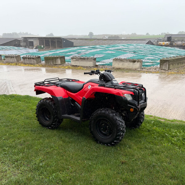 Red ATV parked on wet grass with rain falling and industrial buildings in the background.