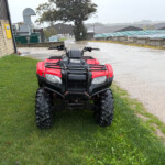 Front view of a red Honda ATV parked on a grassy patch beside a wet concrete path in an industrial yard during rain.