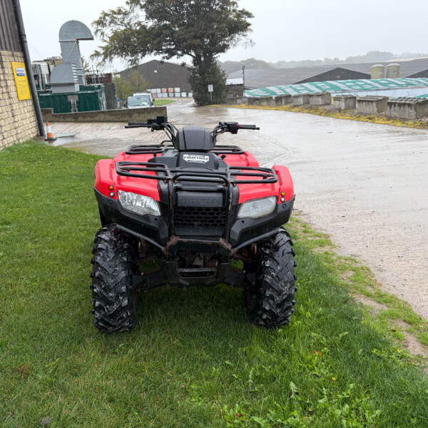 Front view of a red Honda ATV parked on a grassy patch beside a wet concrete path in an industrial yard during rain.