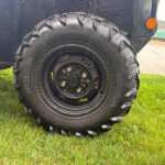 Close-up of a heavy off-road tire on a black rim, with grass beneath and part of the vehicle’s body and an orange marker light visible.