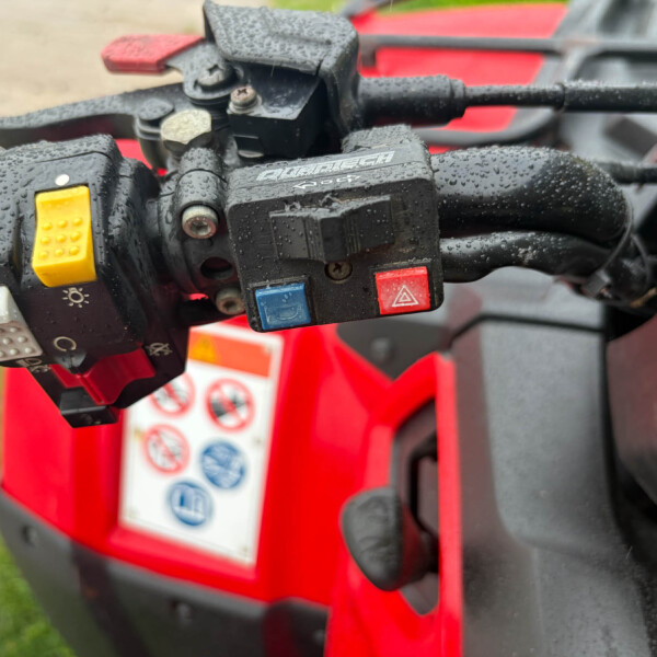 Close-up of red ATV/quad bike handlebar controls with yellow button and blue/red push buttons, wet from rain.