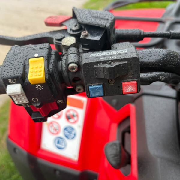 Close-up of rain-dampened motorcycle handlebar controls on a red bike, showing a yellow switch, blue button, and hazard indicators.
