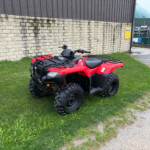 Red ATV parked on a grassy patch beside a stone wall and industrial buildings in the background.