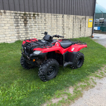 Red ATV parked on a grassy patch beside a stone wall and industrial buildings in the background.