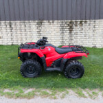 Red ATV parked on the grass in front of a stone wall and dark wooden siding building.