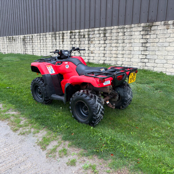 Red four-wheeled ATV parked on grass near a stone wall and a dark metal building in the background.