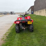 Red ATV/quad bike parked on a grassy verge beside a wide, wet industrial yard with trucks and warehouse buildings in the background.