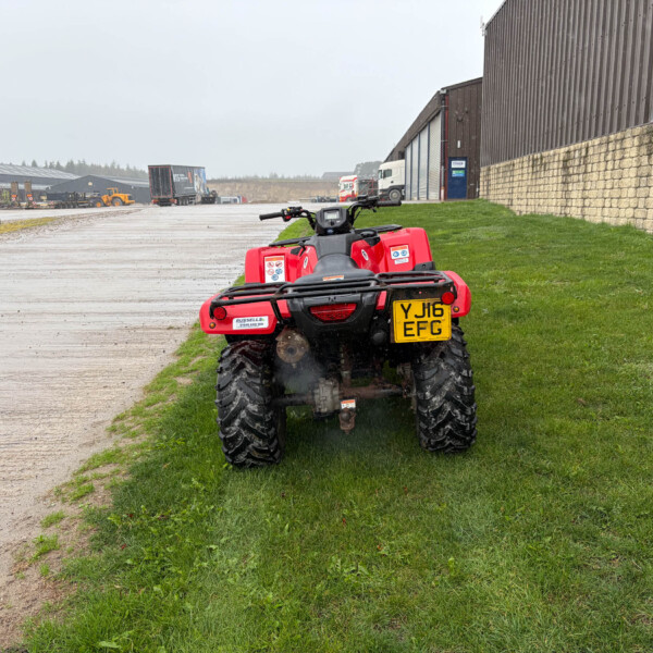 Red ATV/quad bike parked on a grassy verge beside a wide, wet industrial yard with trucks and warehouse buildings in the background.