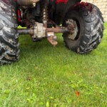 Close-up of a rusty tractor rear axle and hitch assembly between large muddy tires on a grassy lawn, with a stone wall in the background