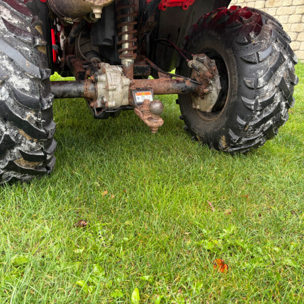 Close-up of a rusty tractor rear axle and hitch assembly between large muddy tires on a grassy lawn, with a stone wall in the background