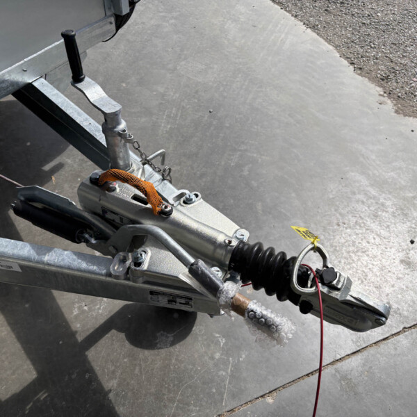 Close-up of a trailer hitch and coupler on a metal trailer frame, with an orange strap and safety chain on a concrete surface.
