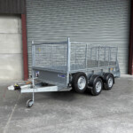 Metal utility trailer with mesh sides and dual axles parked in front of a large corrugated metal roll-up door at an industrial site a yellow door on the right side faintly visible