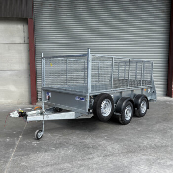 Metal utility trailer with mesh sides and dual axles parked in front of a large corrugated metal roll-up door at an industrial site a yellow door on the right side faintly visible