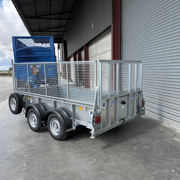 Flatbed trailer with a blue metal cage cargo container parked beside a corrugated warehouse with a large roller door.