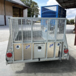 Rear view of a galvanized metal utility trailer with mesh sides and red tail lights in an industrial yard with a blue container in the background.