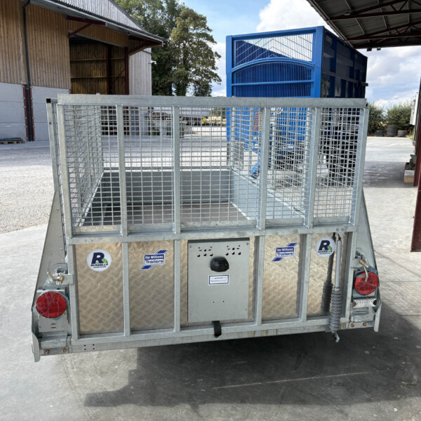 Rear view of a galvanized metal utility trailer with mesh sides and red tail lights in an industrial yard with a blue container in the background.