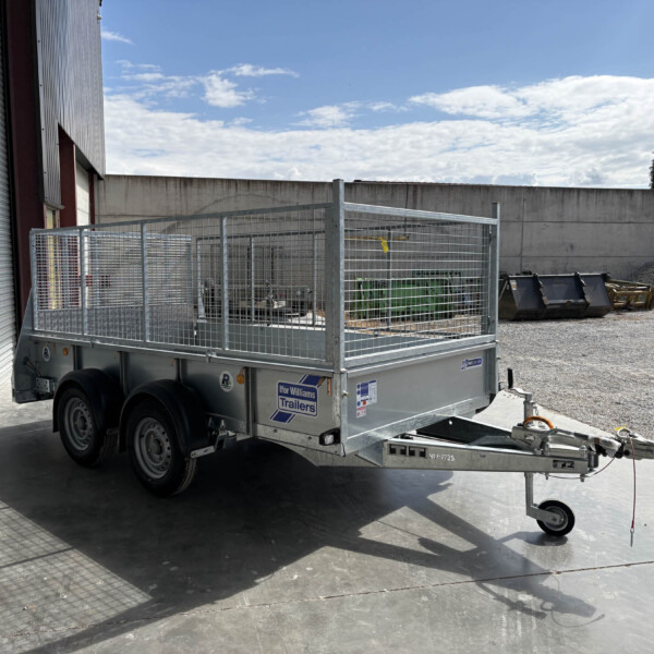 Silver metal utility trailer with mesh sides and a front hitch, parked beside a corrugated metal wall in an industrial yard, sunny day.