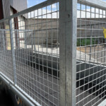 Industrial galvanized wire fence panels forming a gate at an outdoor warehouse area with a yellow tag attached to the mesh.