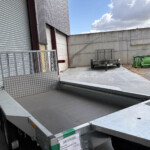 Industrial loading area with a large flatbed trailer, gray ramp, and corrugated metal building under a blue sky with clouds.