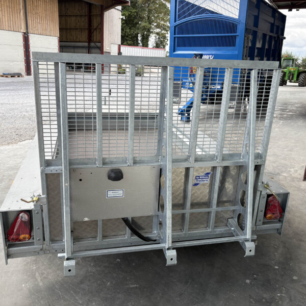 Rear view of a galvanized metal mesh trailer with red taillights in a warehouse yard, blue equipment and a green tractor visible in the background.