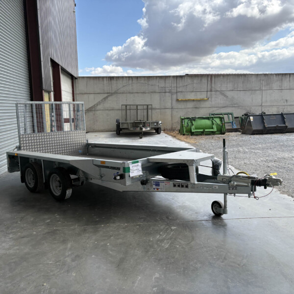 Industrial outdoor area with a metal-clad building, a flatbed trailer, and a tall concrete wall under a cloudy sky, with dumpsters nearby.