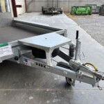 Close-up of a metal utility trailer with a spare tire and hitch, parked on a concrete surface in an industrial yard with dumpsters in the background.