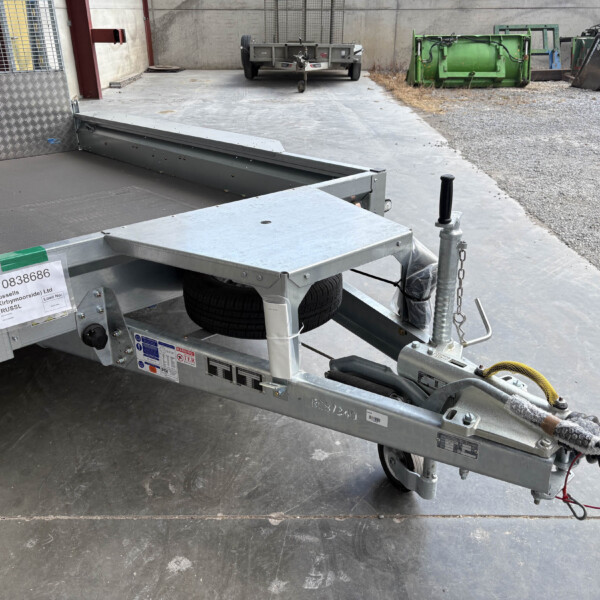 Close-up of a metal utility trailer with a spare tire and hitch, parked on a concrete surface in an industrial yard with dumpsters in the background.