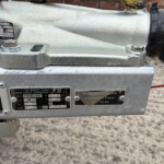 Close-up of a silver trailer hitch or coupling mechanism with bolts and metal plates, showing maker labels on a gray surface.