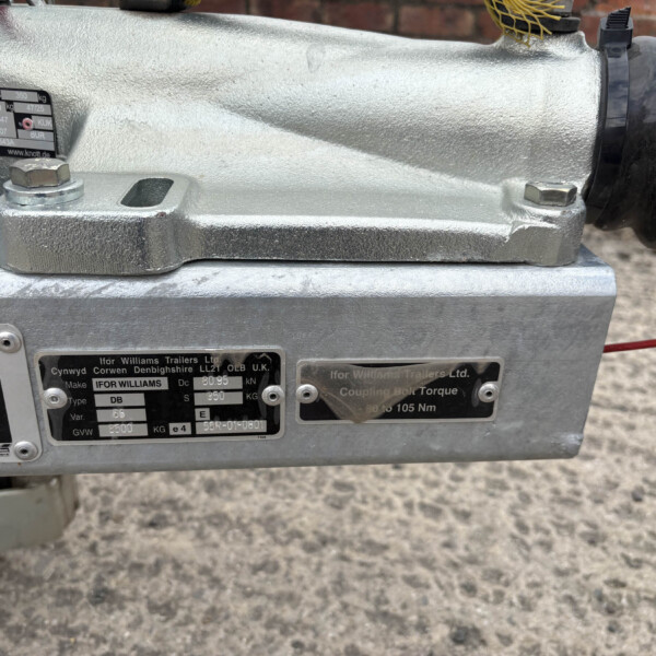 Close-up of a silver trailer hitch or coupling mechanism with bolts and metal plates, showing maker labels on a gray surface.