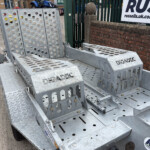 Close-up of a rugged galvanized trailer step with perforated treads and side handles, parked beside a brick wall.