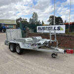 Silver metal trailer with multiple axles and perforated sides parked in an industrial yard, with a blue tractor and Russell's sign in the background behind a green fence.