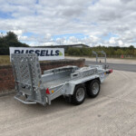 Metal flatbed trailer with perforated side rails beside a brick wall and a large 'RUSSELLS' sign on a cloudy day.