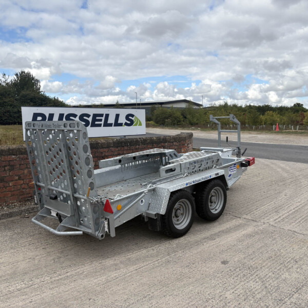 Metal flatbed trailer with perforated side rails beside a brick wall and a large 'RUSSELLS' sign on a cloudy day.