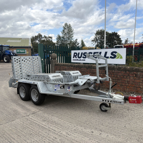 Silver metal trailer with multiple axles and perforated sides parked in an industrial yard, with a blue tractor and Russell's sign in the background behind a green fence.