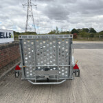 Back of a perforated metal Ifor Williams trailer with timber gate, parked by a brick wall and Russell’s sign in a rural lot.