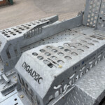 Close-up of a perforated galvanized steel trailer step with bolts and cut-out holes for grip and drainage, partially showing branding on the side panels.