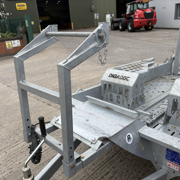 Close-up view of a metal trailer frame with a hanging chain, set in an industrial yard with a workshop and a red vehicle in the background.
