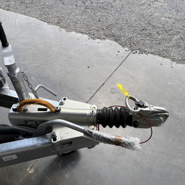 Close-up of a trailer hitch assembly with a vertical black handle, safety chain, and yellow tag on a concrete surface.