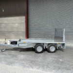 Industrial trailer with a spare tire and towing hitch parked on a concrete lot beside a large corrugated metal wall and roll-up door.