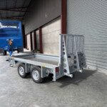 Blue livestock trailer attached to a gray flatbed trailer in an industrial loading area beside a corrugated metal warehouse wall with a roll-up door.
