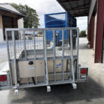 Metal trailer with a mesh cage in a loading yard between shed-style buildings and a blue industrial machine.