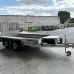 Silver flatbed trailer parked in a loading yard beside metal cages and dumpsters against a long gray concrete wall.