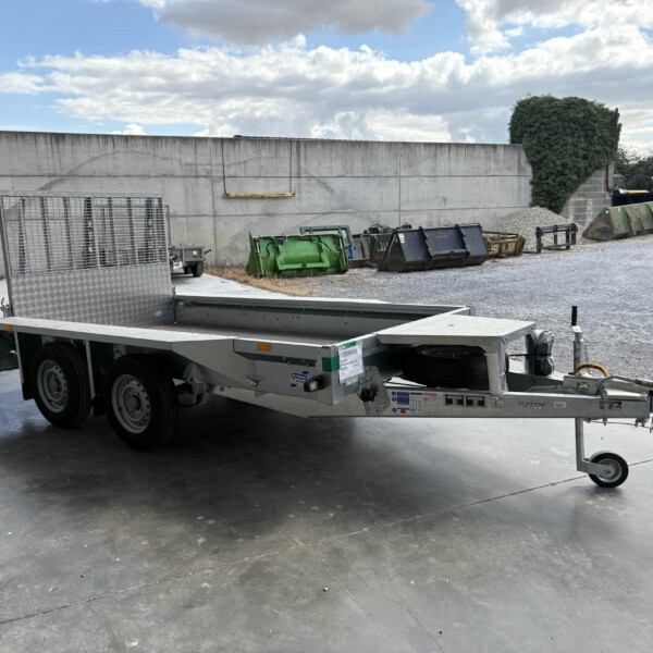 Silver flatbed trailer parked in a loading yard beside metal cages and dumpsters against a long gray concrete wall.
