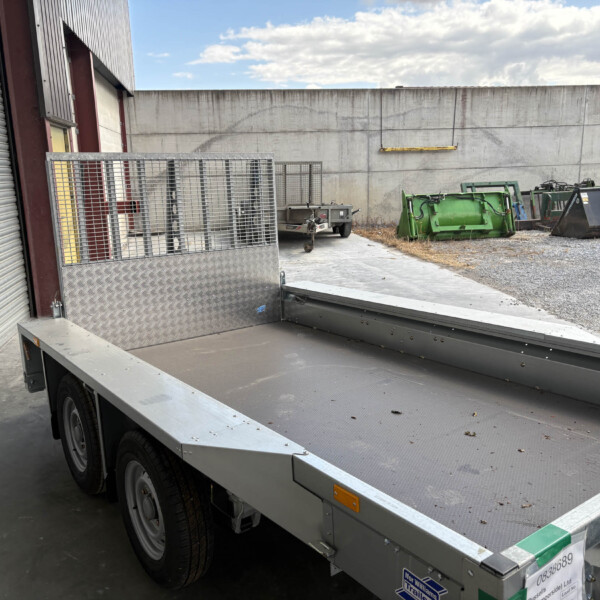 Industrial loading yard with a metal flatbed trailer and ramp, parked beside a corrugated metal building and a concrete wall.