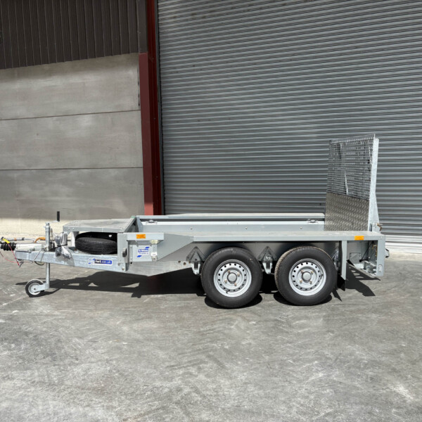 Silver utility trailer with spare tire and diamond-plate ramp, parked near a corrugated metal wall in an industrial area.